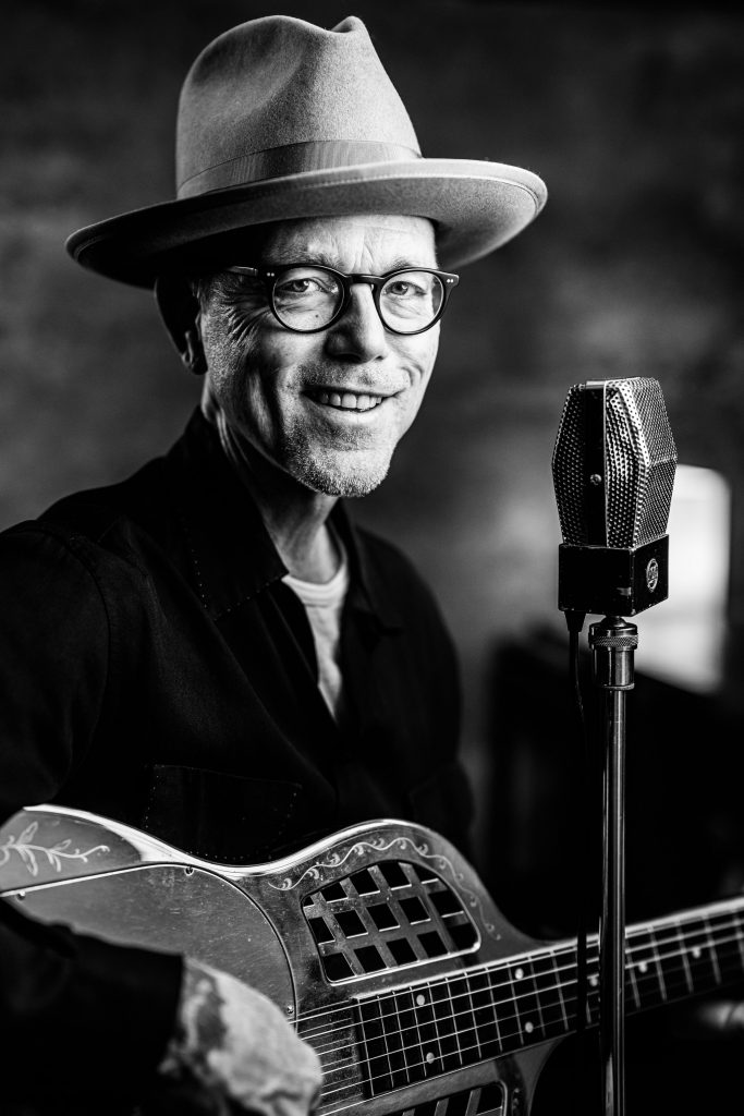 Pete smiles into the camera with his trademark stetson hat and tri-cone resonator guitar.